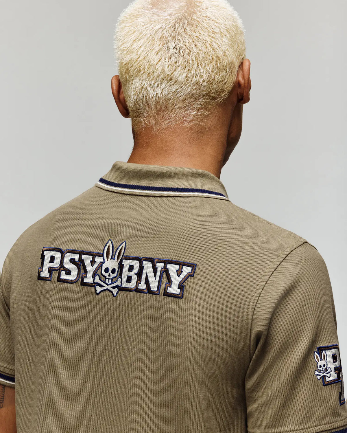 A person with short, bleached blond hair is seen from behind wearing a Psycho Bunny MENS WADE PATCH PIQUE POLO SHIRT (MT0100109) in olive green, featuring “PSYBNY” and an embroidered Bunny skull logo on the back and sleeve patch.