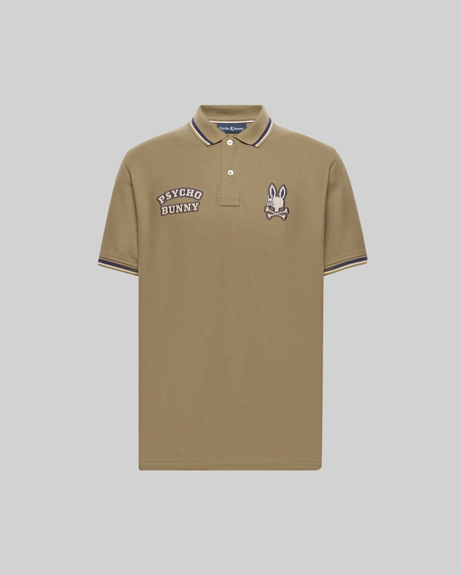 The Psycho Bunny Men's Wade Patch Pique Polo Shirt (MT0100109) is made from khaki Pima cotton with white buttons, "PSYCHO BUNNY" text, an embroidered Bunny skull logo patch on the chest, and white/navy striped collar and sleeve trim.