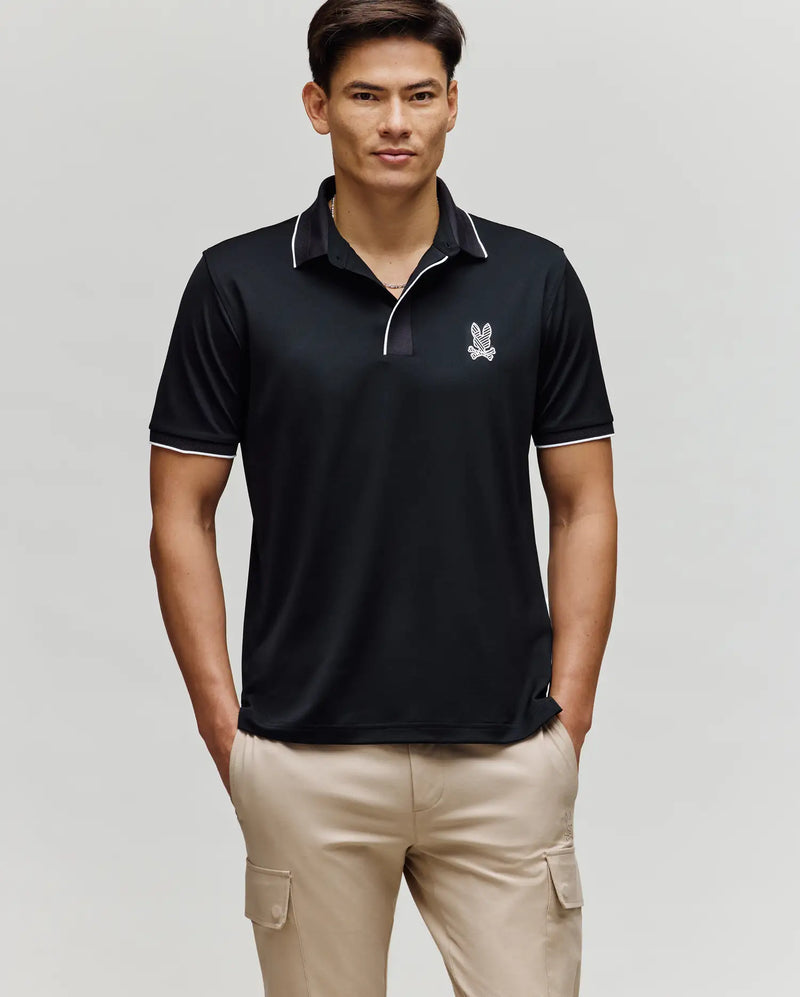 A man stands facing forward wearing the Psycho Bunny MENS JAYDEN SPORT POLO - MT0100111 in black with white trim and a small bunny logo on the chest, paired with light beige cargo pants. He has his hands in his pockets and a neutral expression.