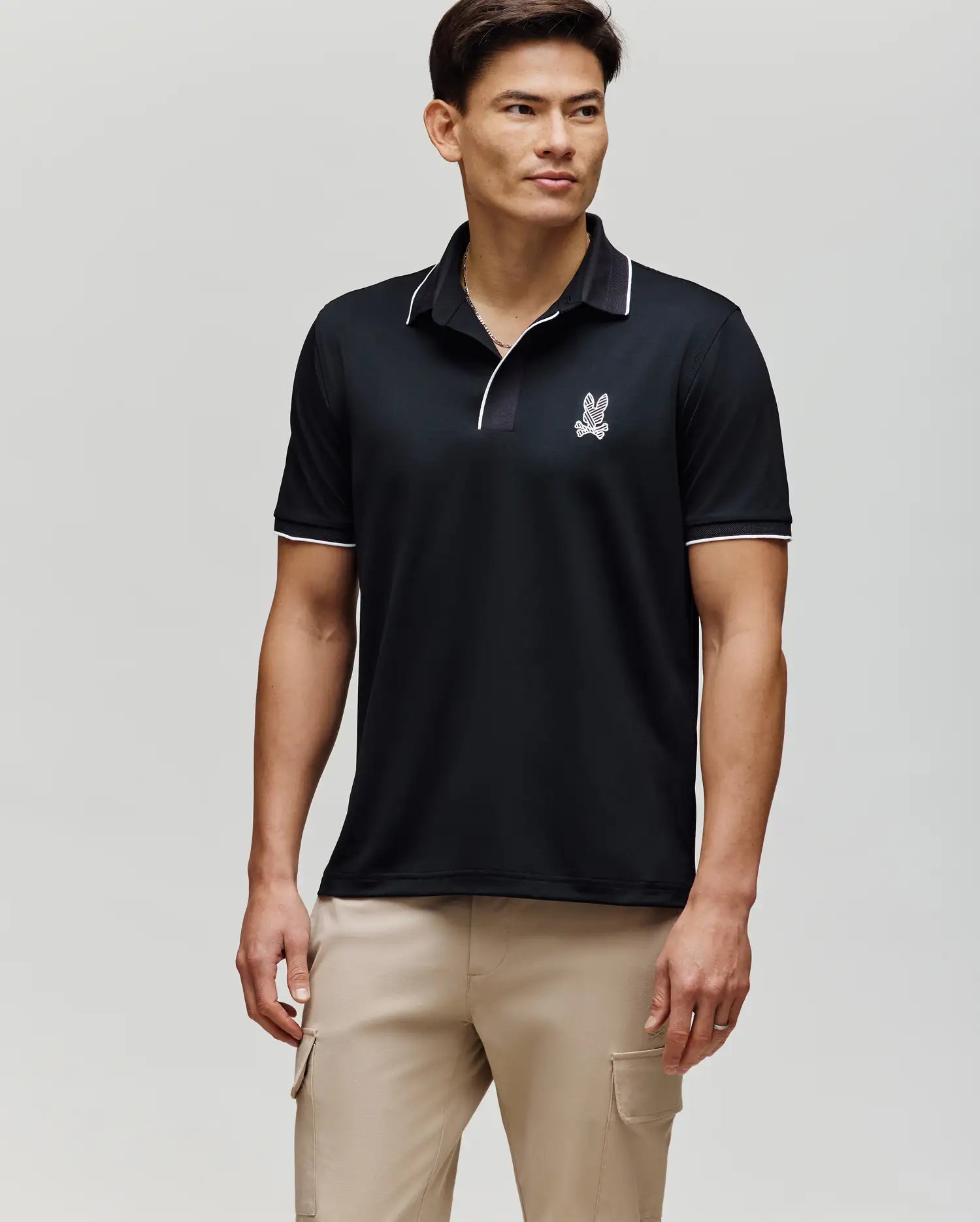 A man with short dark hair stands facing forward in a Psycho Bunny MENS JAYDEN SPORT POLO (MT0100111), featuring a black athletic fit with white trim and a small logo on the chest, paired with light beige cargo pants against a light gray background.