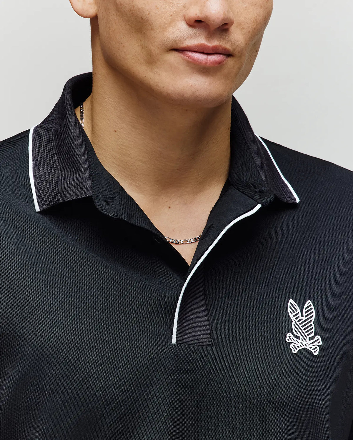 A person wears the Psycho Bunny MENS JAYDEN SPORT POLO - MT0100111 in black with white trim and an embroidered bunny logo on the left chest; moisture-wicking fabric ensures comfort, showing only the lower face and upper torso.