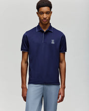 A man stands against a plain background wearing light gray pants and a Psycho Bunny MENS JAYDEN SPORT POLO - MT0100111 in navy blue, featuring a white chest logo. Its athletic fit and moisture-wicking fabric offer style and comfort.