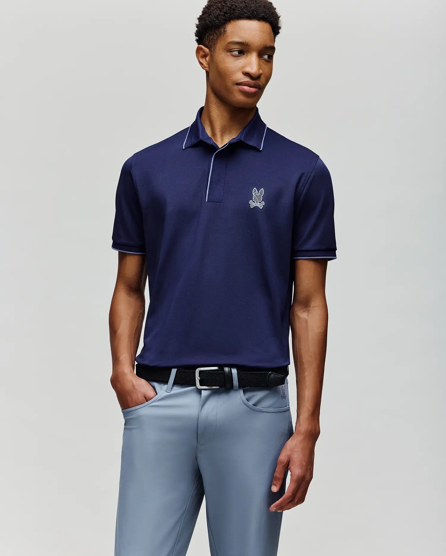 A man wears the Psycho Bunny MENS JAYDEN SPORT POLO - MT0100111 in dark navy with a white logo and athletic fit, paired with light gray trousers, standing against a plain light background with one hand in his pocket.