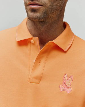 Mens Olivier Pique Polo in vibrant peach, featuring a classic fit, two-button placket, and a small embroidered pink and cream Psycho Bunny skull logo on the left chest, with subtle pink tipping on the collar.