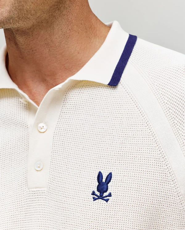 A close-up of a person wearing the Psycho Bunny MENS LAZARE KNIT POLO (MT0100165), an off-white textured knit with a blue collar trim and a blue embroidered Psycho Bunny logo on the chest.