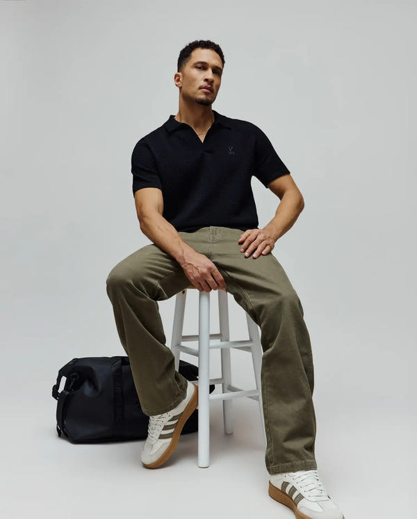 Mens Devon Knit Polo, a black textured knit polo shirt with an open collar and a small embroidered Psycho Bunny skull logo on the left chest, offering a classic fit.