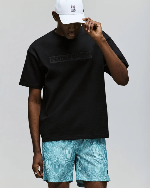 A person wearing the Psycho Bunny MENS ZAYN OVERSIZED GRAPHIC TEE (MT0200004), made from 100% cotton, paired with light blue bunny-patterned shorts and a white cap featuring a small bunny logo, stands against a plain background.