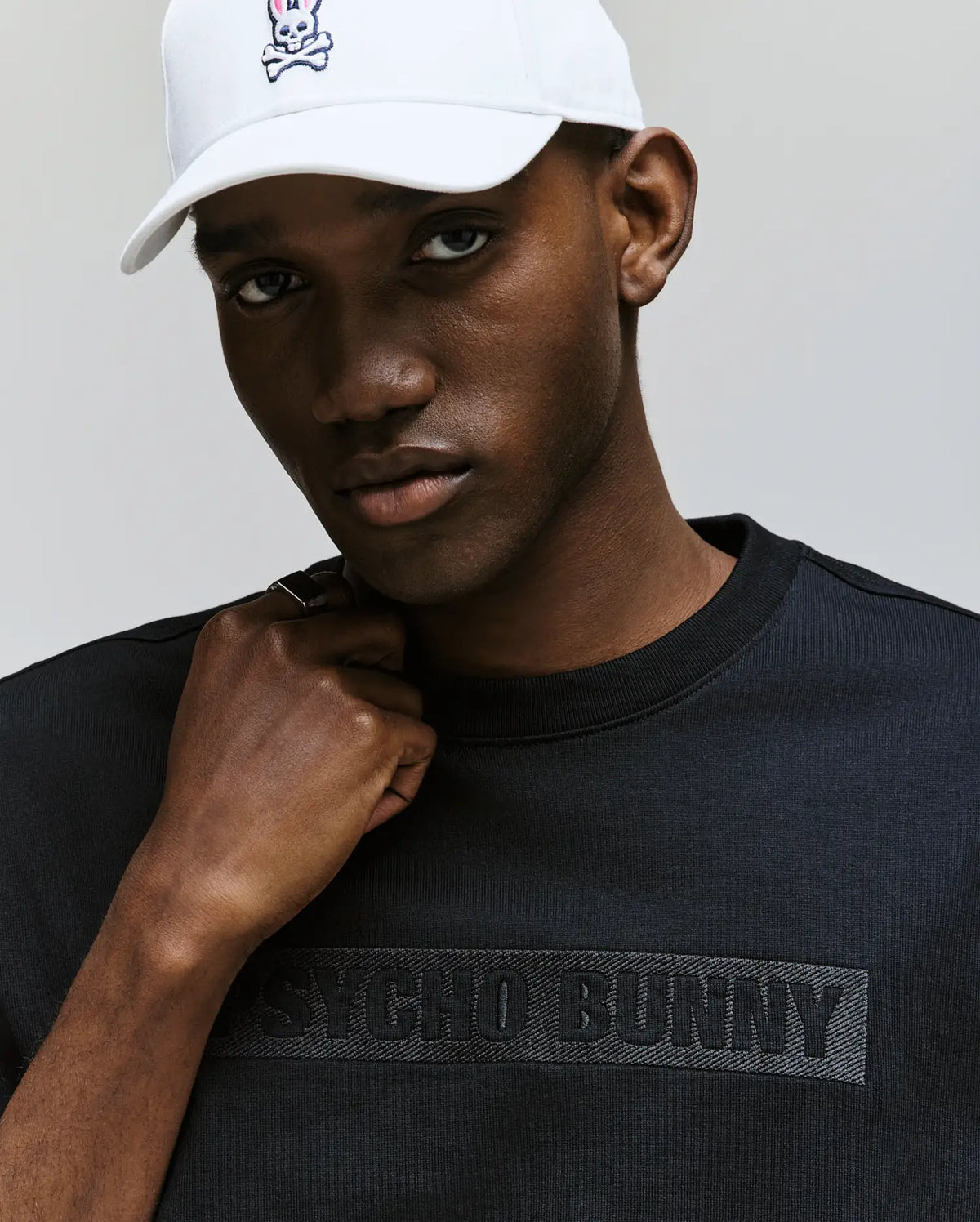 A man wearing the Psycho Bunny MENS ZAYN OVERSIZED GRAPHIC TEE - MT0200004 and a white cap with a cartoon bunny logo looks at the camera with a neutral expression.