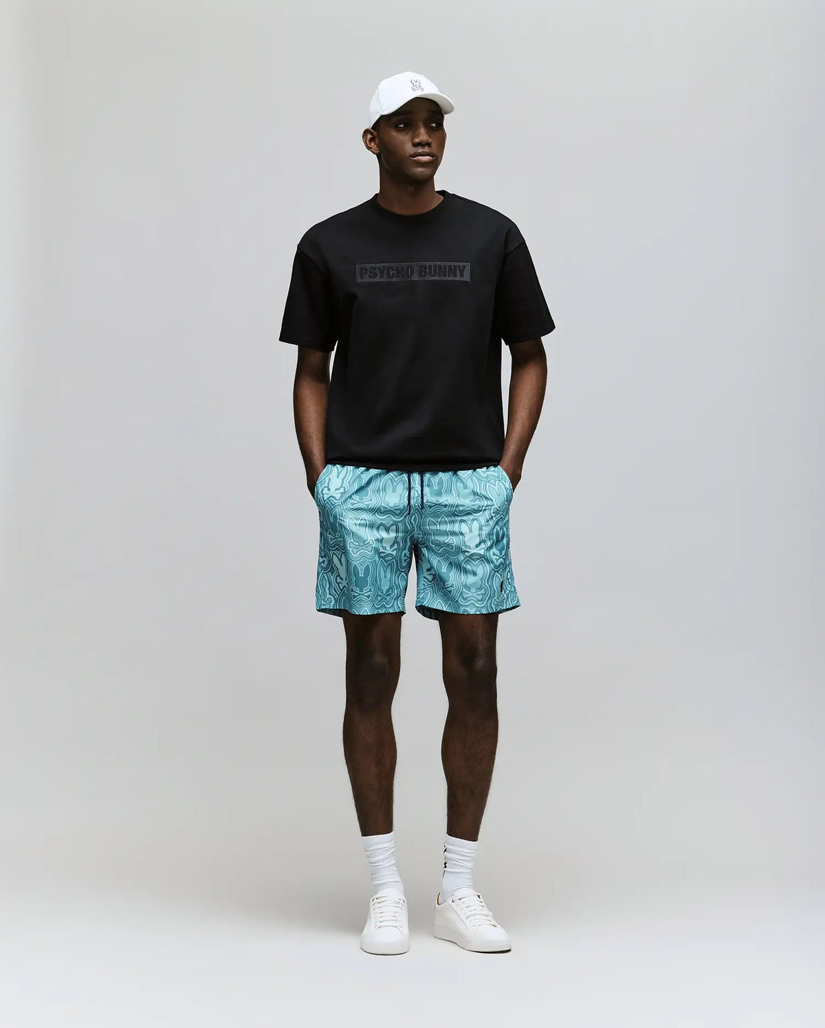 A man stands against a plain background wearing the Psycho Bunny MENS ZAYN OVERSIZED GRAPHIC TEE - MT0200004 in black, paired with turquoise patterned shorts, a white cap, white socks, and white sneakers, hands in his shorts pockets.