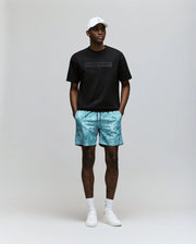 A man stands against a plain background wearing the Psycho Bunny MENS ZAYN OVERSIZED GRAPHIC TEE - MT0200004 in black, paired with turquoise patterned shorts, a white cap, white socks, and white sneakers, hands in his shorts pockets.