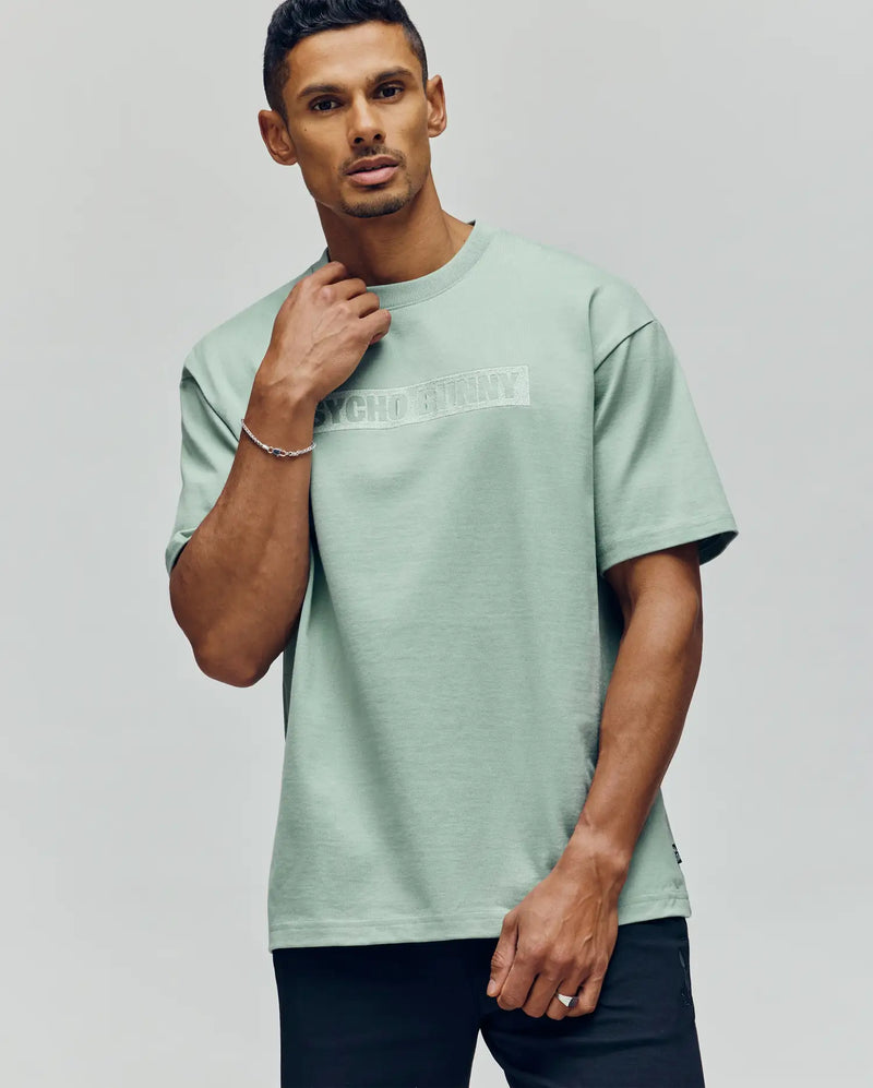 A man wears the Psycho Bunny MENS ZAYN OVERSIZED GRAPHIC TEE (MT0200004), a light green heavyweight jersey with a relaxed fit and "PSYCHO" partially visible on the chest, standing against a plain background.