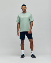 A man stands against a plain background wearing the Psycho Bunny MENS ZAYN OVERSIZED GRAPHIC TEE - MT0200004 in light green, paired with black shorts, white socks, and black sneakers. He faces forward with a neutral expression and relaxed posture.