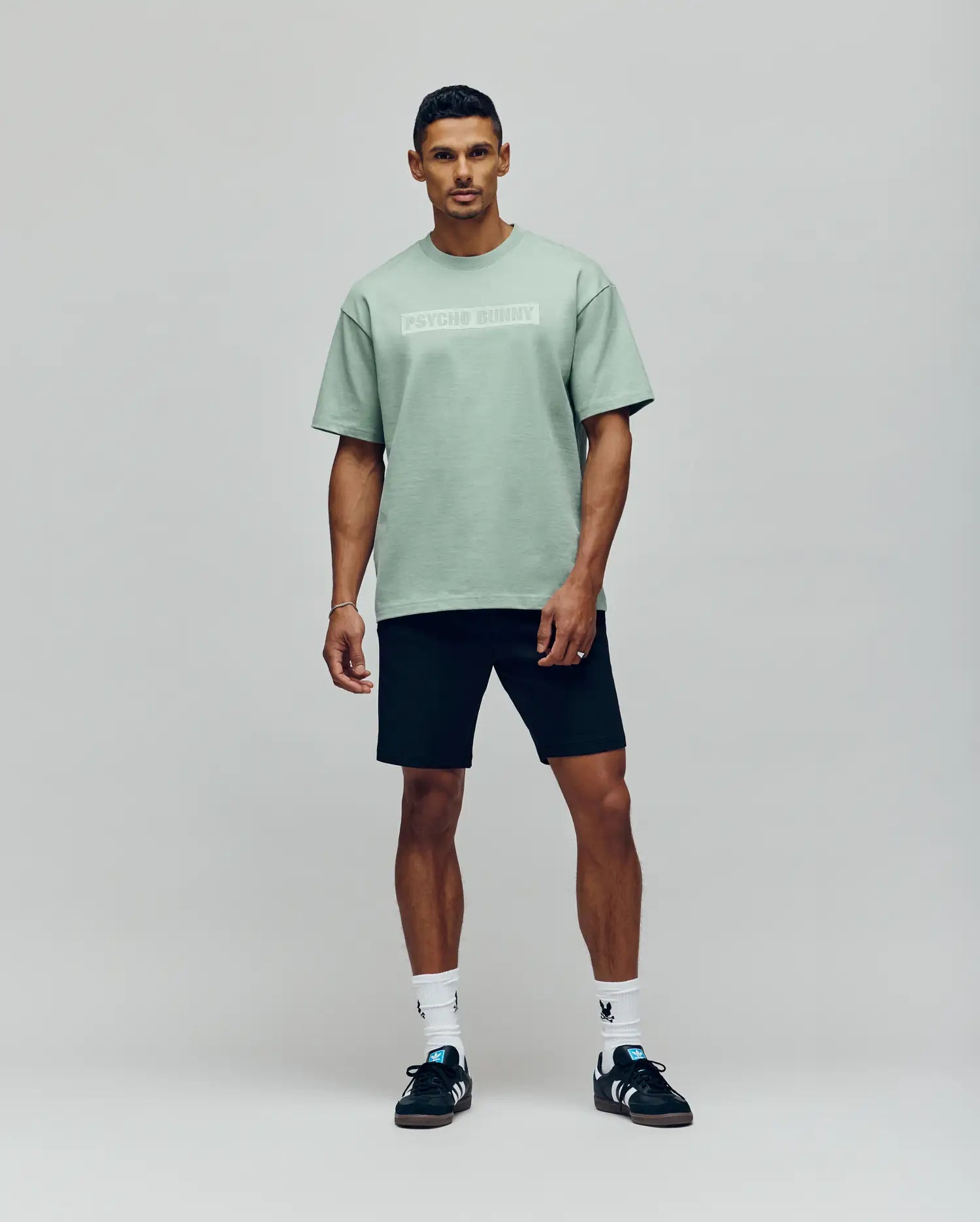 A man stands against a plain background wearing the Psycho Bunny MENS ZAYN OVERSIZED GRAPHIC TEE - MT0200004 in light green, paired with black shorts, white socks, and black sneakers. He faces forward with a neutral expression and relaxed posture.