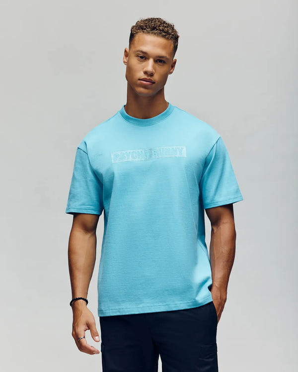 A young man wears the Psycho Bunny MENS ZAYN OVERSIZED GRAPHIC TEE - MT0200004 in light blue with subtle white text and black pants, standing confidently against a plain light gray background.