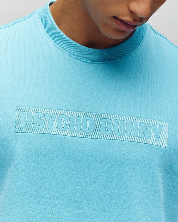 A man in a relaxed fit, light blue MENS ZAYN OVERSIZED GRAPHIC TEE by Psycho Bunny, featuring tonal "PSYCHO BUNNY" chest embroidery. Only his lower face, neck, and upper torso are visible.