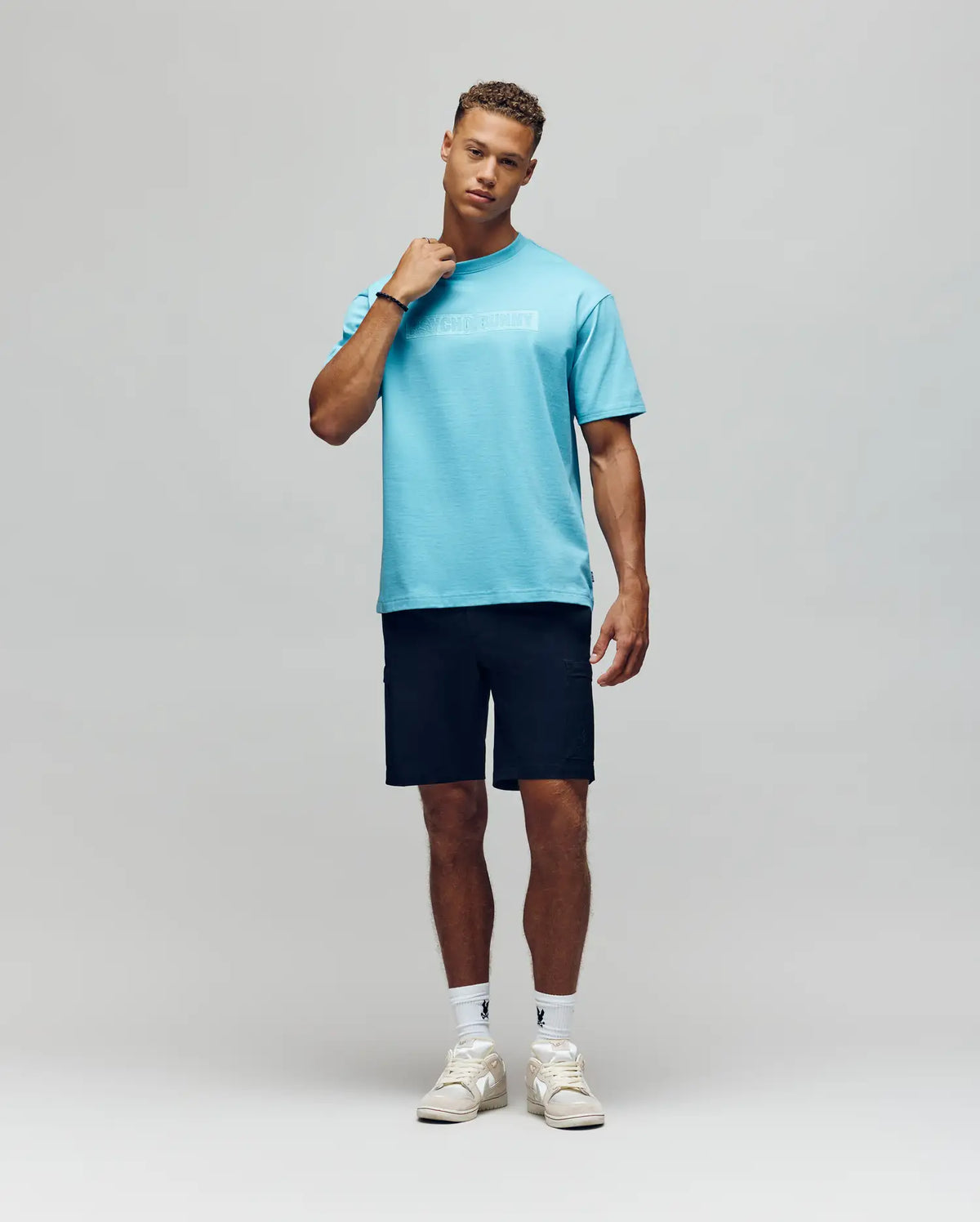 A man poses against a plain background wearing the Psycho Bunny MENS ZAYN OVERSIZED GRAPHIC TEE (MT0200004), paired with black shorts, white socks, and white sneakers. He touches his neck and has a relaxed expression.