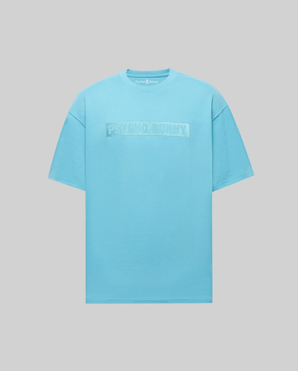 The Psycho Bunny MENS ZAYN OVERSIZED GRAPHIC TEE (MT0200004) features a relaxed fit, light blue color, short sleeves, and the embroidered wordmark logo in matching blue across the chest against a gray background.
