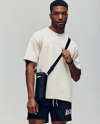 A man wears a Psycho Bunny MENS ZAYN OVERSIZED GRAPHIC TEE (MT0200004) in cream and black shorts, holding a black water bottle with a strap. The relaxed fit stands out against the plain, light background.