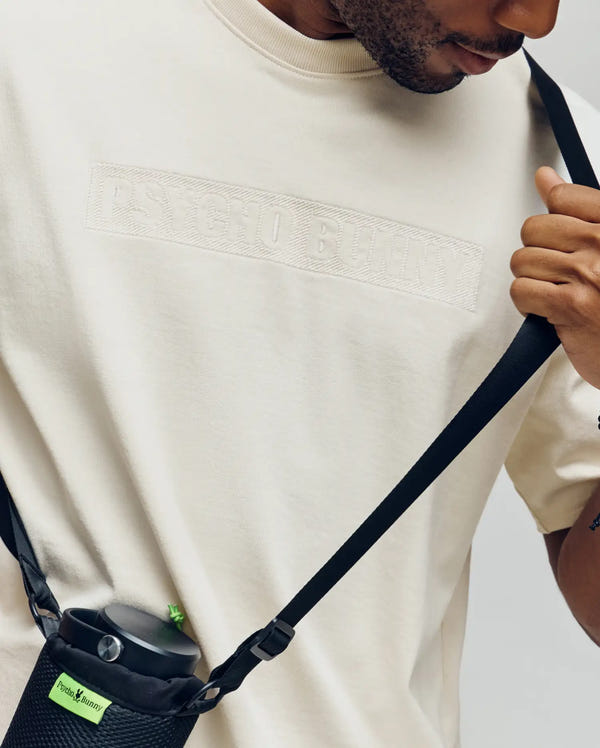 A person in a Psycho Bunny MENS ZAYN OVERSIZED GRAPHIC TEE (MT0200004) holds a black crossbody strap connected to a black water bottle holder with a green Psycho Bunny tag.