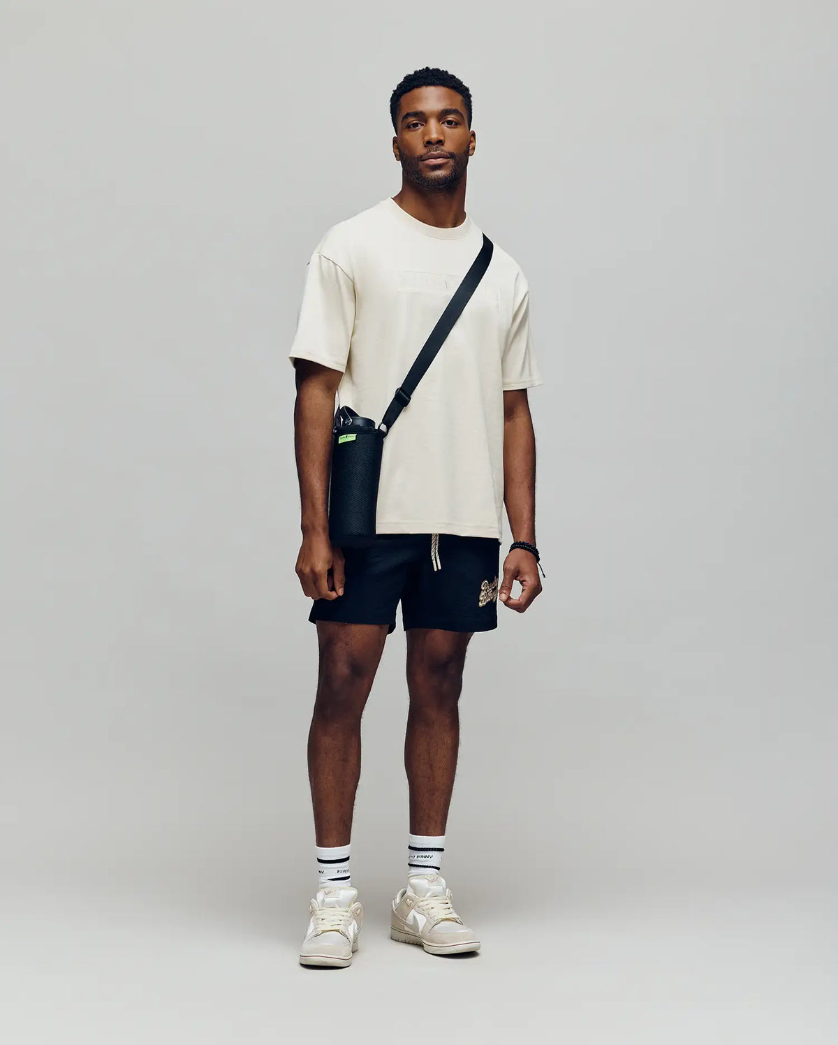 A man stands against a plain background, wearing a Psycho Bunny MENS ZAYN OVERSIZED GRAPHIC TEE - MT0200004 in beige, with black shorts, white socks, beige sneakers, and a black crossbody bag. He faces the camera with a neutral expression.