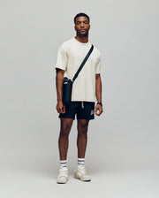 A man stands against a plain background, wearing a Psycho Bunny MENS ZAYN OVERSIZED GRAPHIC TEE - MT0200004 in beige, with black shorts, white socks, beige sneakers, and a black crossbody bag. He faces the camera with a neutral expression.
