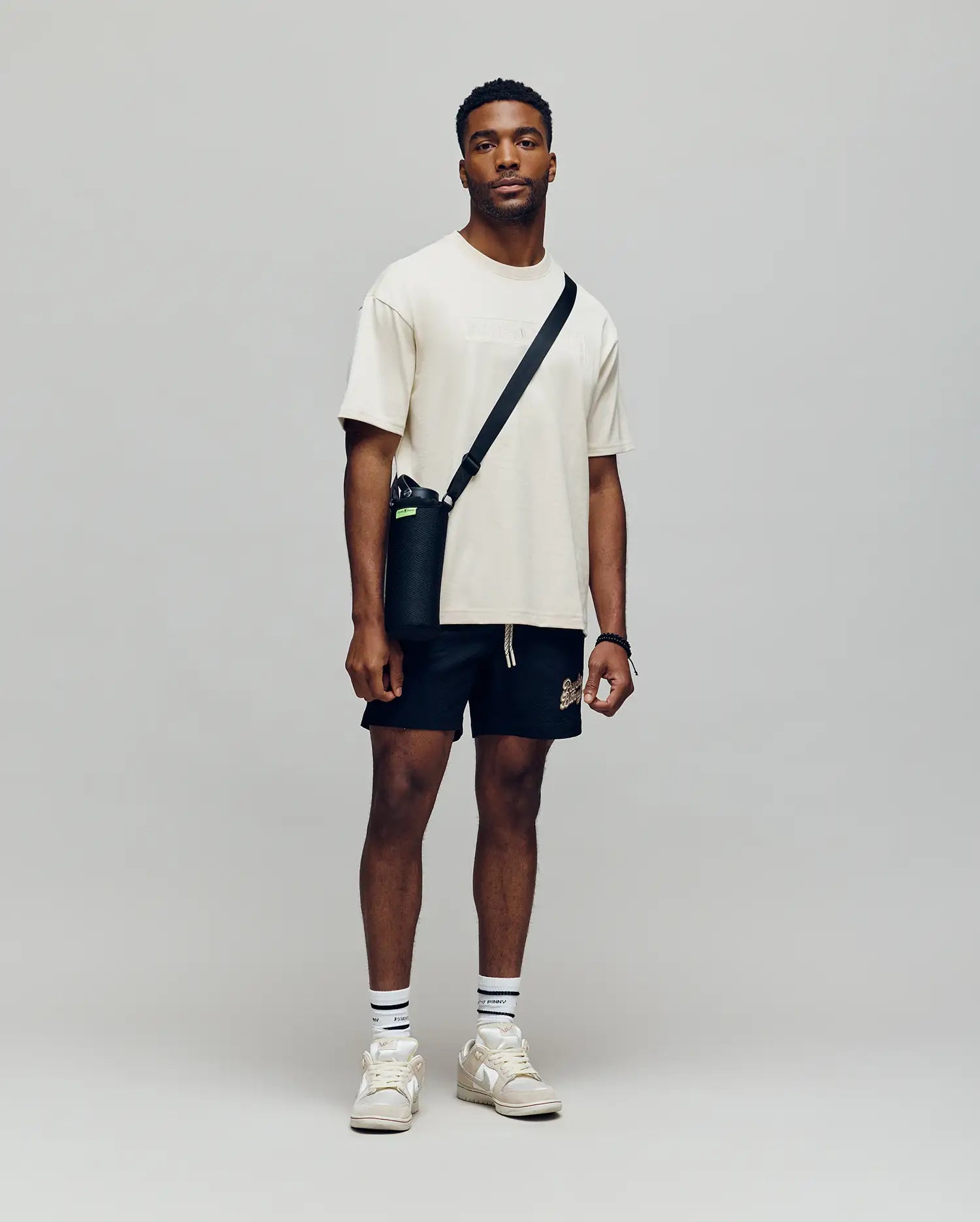 A man stands against a plain background, wearing a Psycho Bunny MENS ZAYN OVERSIZED GRAPHIC TEE - MT0200004 in beige, with black shorts, white socks, beige sneakers, and a black crossbody bag. He faces the camera with a neutral expression.