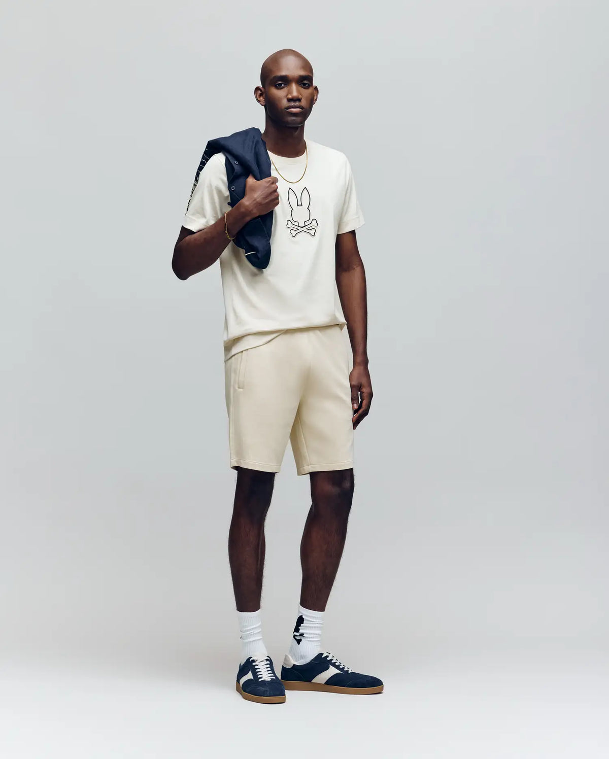 A man stands against a plain background wearing the Psycho Bunny MENS SETH GRAPHIC TEE - MT0200005 in cream, matching shorts, white socks, dark sneakers, and holds a dark jacket over his shoulder.