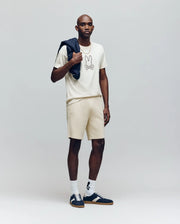 A man stands against a plain background wearing the Psycho Bunny MENS SETH GRAPHIC TEE - MT0200005 in cream, matching shorts, white socks, dark sneakers, and holds a dark jacket over his shoulder.