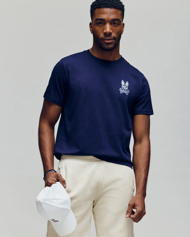 A man stands against a neutral background in Psycho Bunny’s MENS DEREK CHEST GRAPHIC TEE - MT0200009, a navy Pima cotton tee with a small white bunny chest print, cream shorts, and holding a white baseball cap in his left hand.
