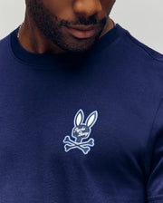 A man wears the Psycho Bunny MENS DEREK CHEST GRAPHIC TEE (MT0200009), a navy blue Pima cotton shirt featuring a small bunny head and crossbones graphic with "Psycho Bunny" on the chest.