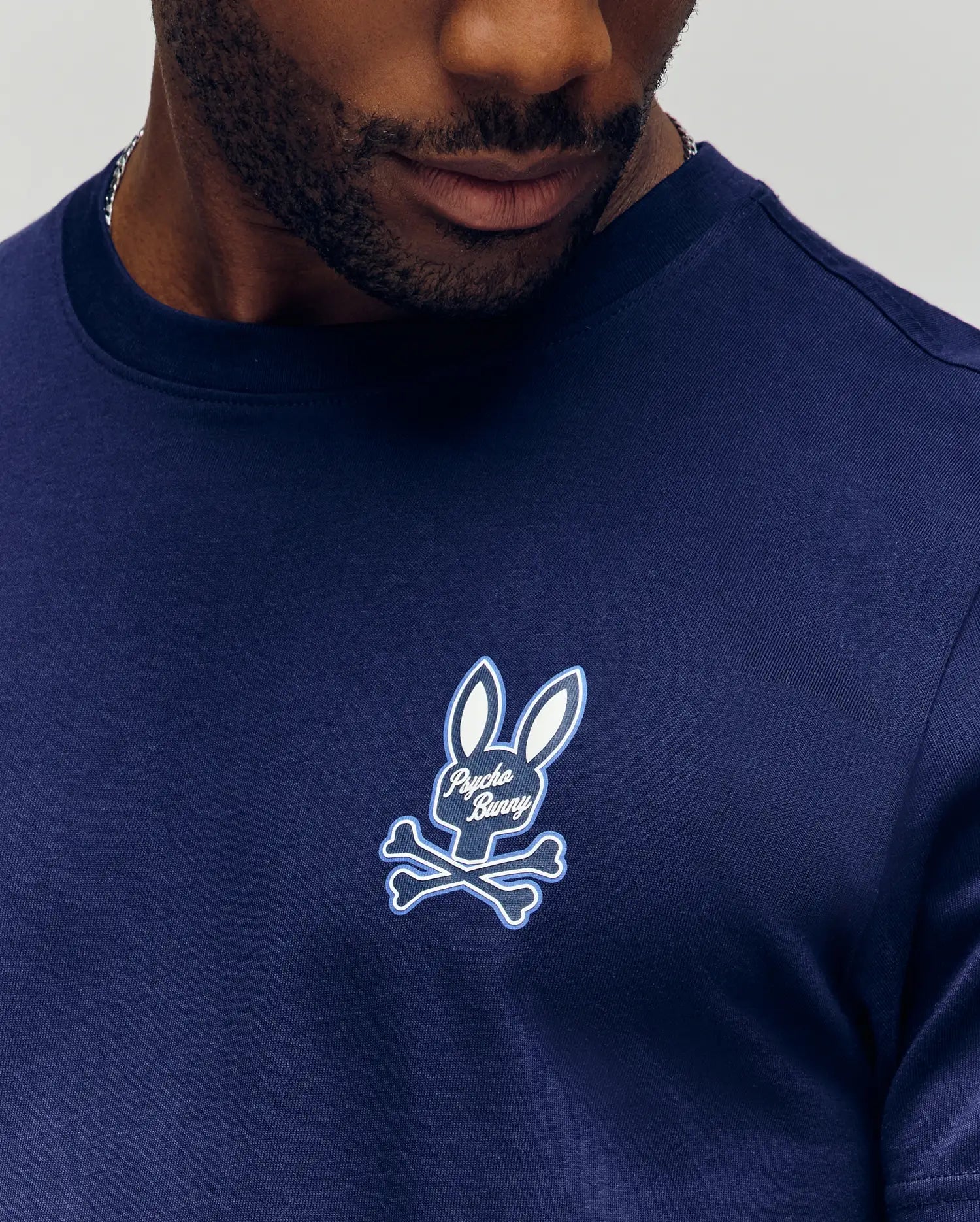A man wears the Psycho Bunny MENS DEREK CHEST GRAPHIC TEE (MT0200009), a navy blue Pima cotton shirt featuring a small bunny head and crossbones graphic with "Psycho Bunny" on the chest.