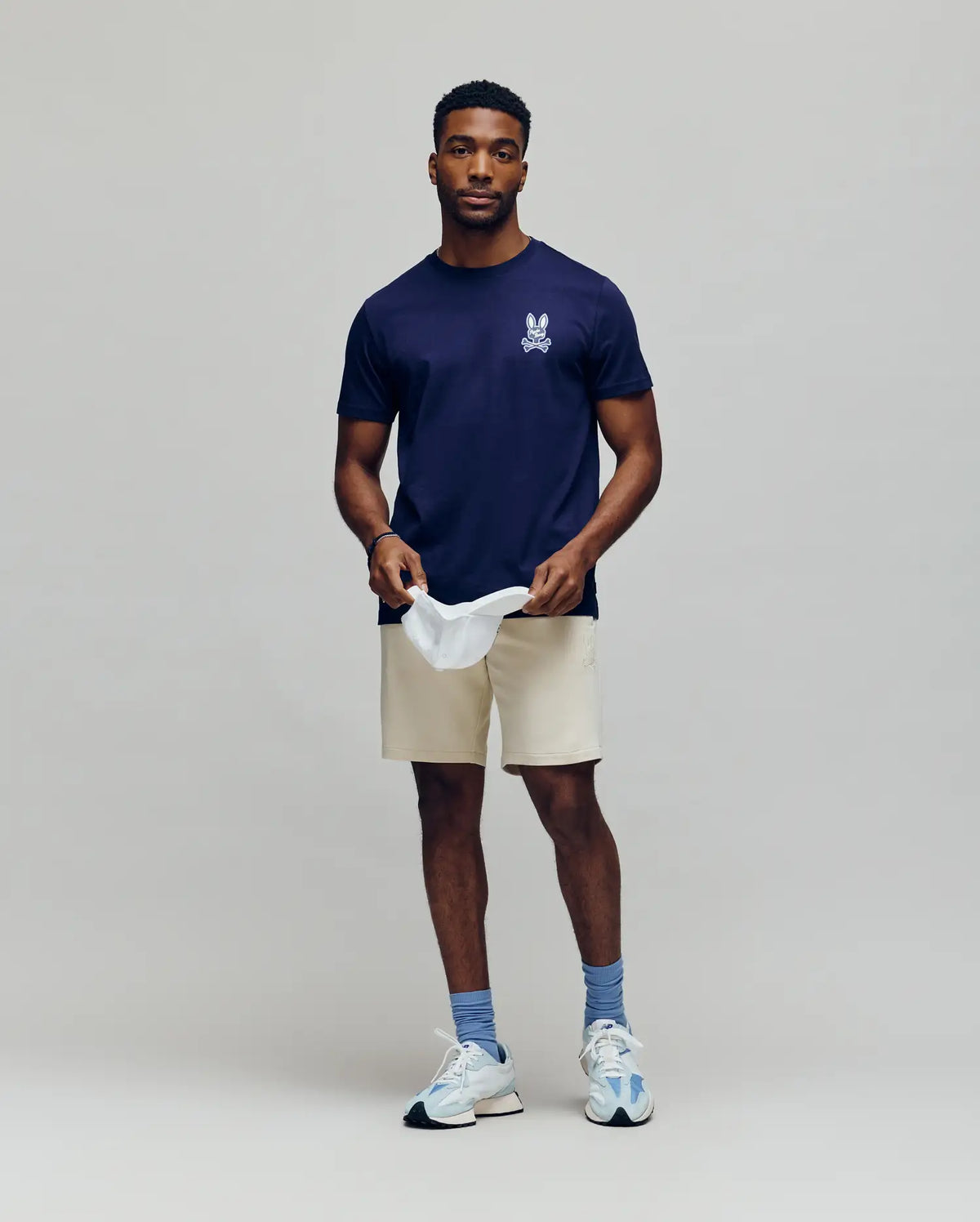A man stands against a plain background, wearing the Psycho Bunny MENS DEREK CHEST GRAPHIC TEE (MT0200009) in navy blue with a small white chest graphic, beige shorts, blue socks, white sneakers, and holding a white cap while looking at the camera.
