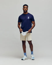 A man stands against a plain background, wearing the Psycho Bunny MENS DEREK CHEST GRAPHIC TEE (MT0200009) in navy blue with a small white chest graphic, beige shorts, blue socks, white sneakers, and holding a white cap while looking at the camera.