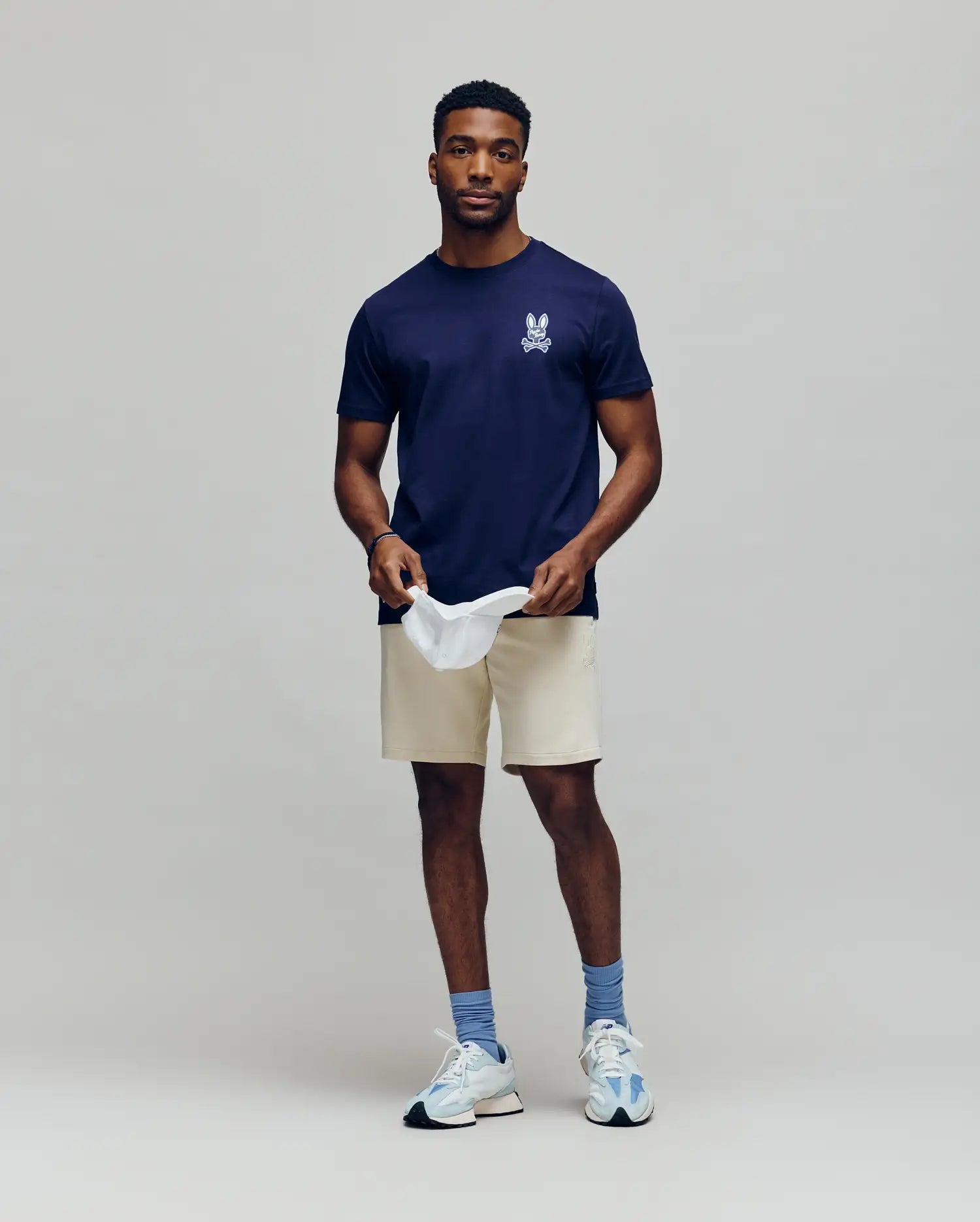 A man stands against a plain background, wearing the Psycho Bunny MENS DEREK CHEST GRAPHIC TEE (MT0200009) in navy blue with a small white chest graphic, beige shorts, blue socks, white sneakers, and holding a white cap while looking at the camera.