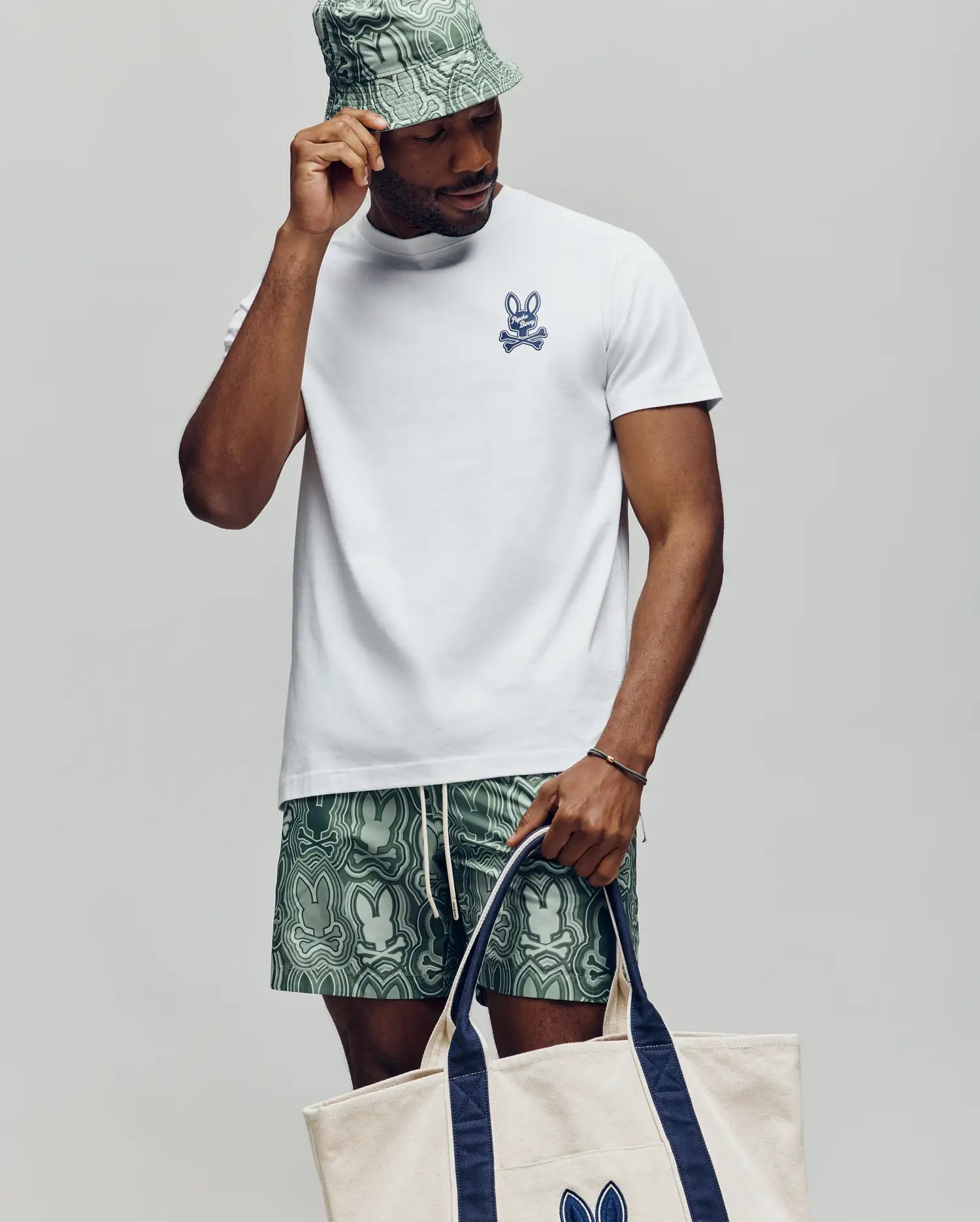 A man wears the Psycho Bunny MENS DEREK CHEST GRAPHIC TEE (MT0200009), green patterned shorts, a matching bucket hat, and carries a beige tote bag with navy handles and Bunny logo, standing against a plain light background.