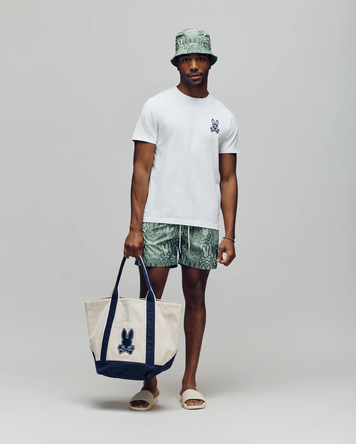 A man stands against a plain background wearing a light green bucket hat, green patterned shorts, beige sandals, and holding a large beige and navy tote bag with a bunny print. He sports Psycho Bunny’s MENS DEREK CHEST GRAPHIC TEE - MT0200009.