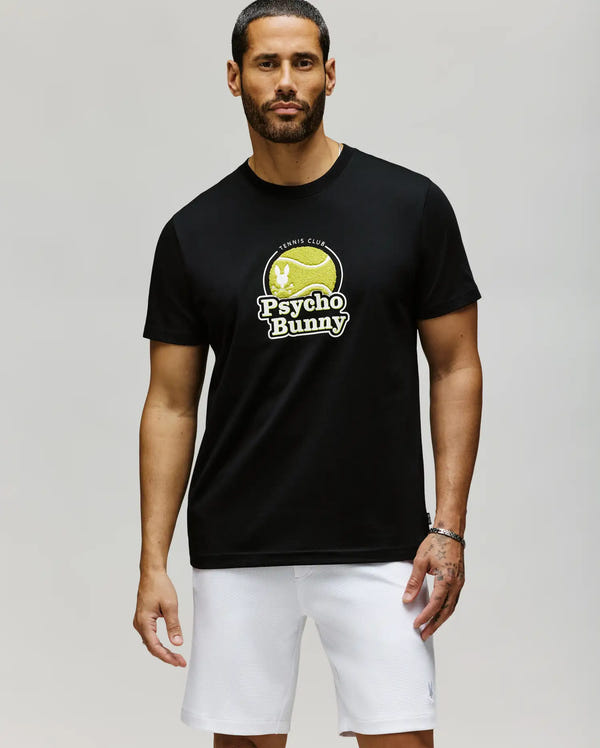 A man with short dark hair and a beard wears the Psycho Bunny MENS PETE GRAPHIC TEE (MT0200012), a black Pima cotton T-shirt with a yellow tennis club logo, paired with white shorts, standing against a light gray background.