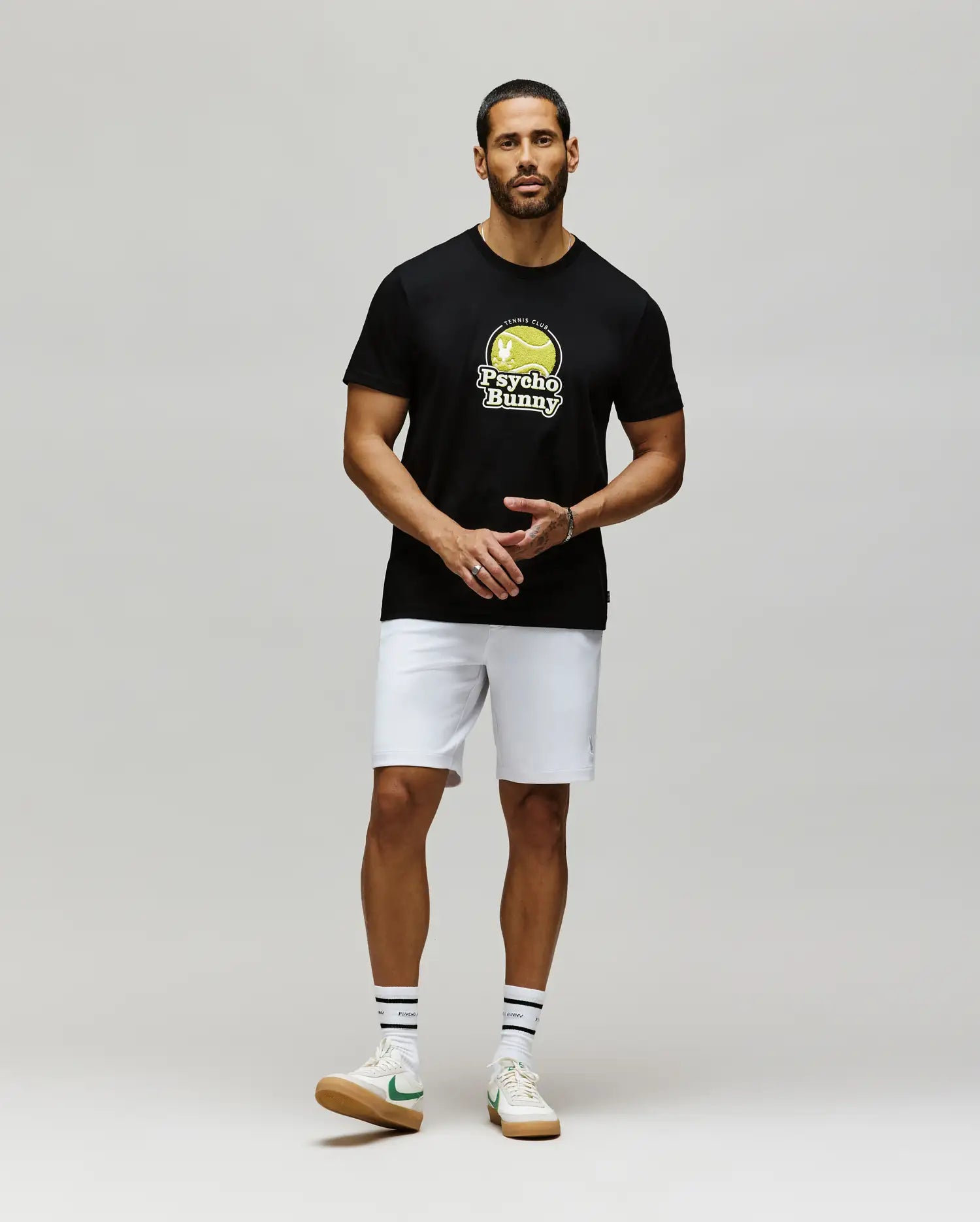 A man stands against a plain background wearing Psycho Bunny's MENS PETE GRAPHIC TEE (MT0200012) in black Pima cotton, paired with white shorts, white tennis socks featuring black stripes, and white sneakers with green accents.
