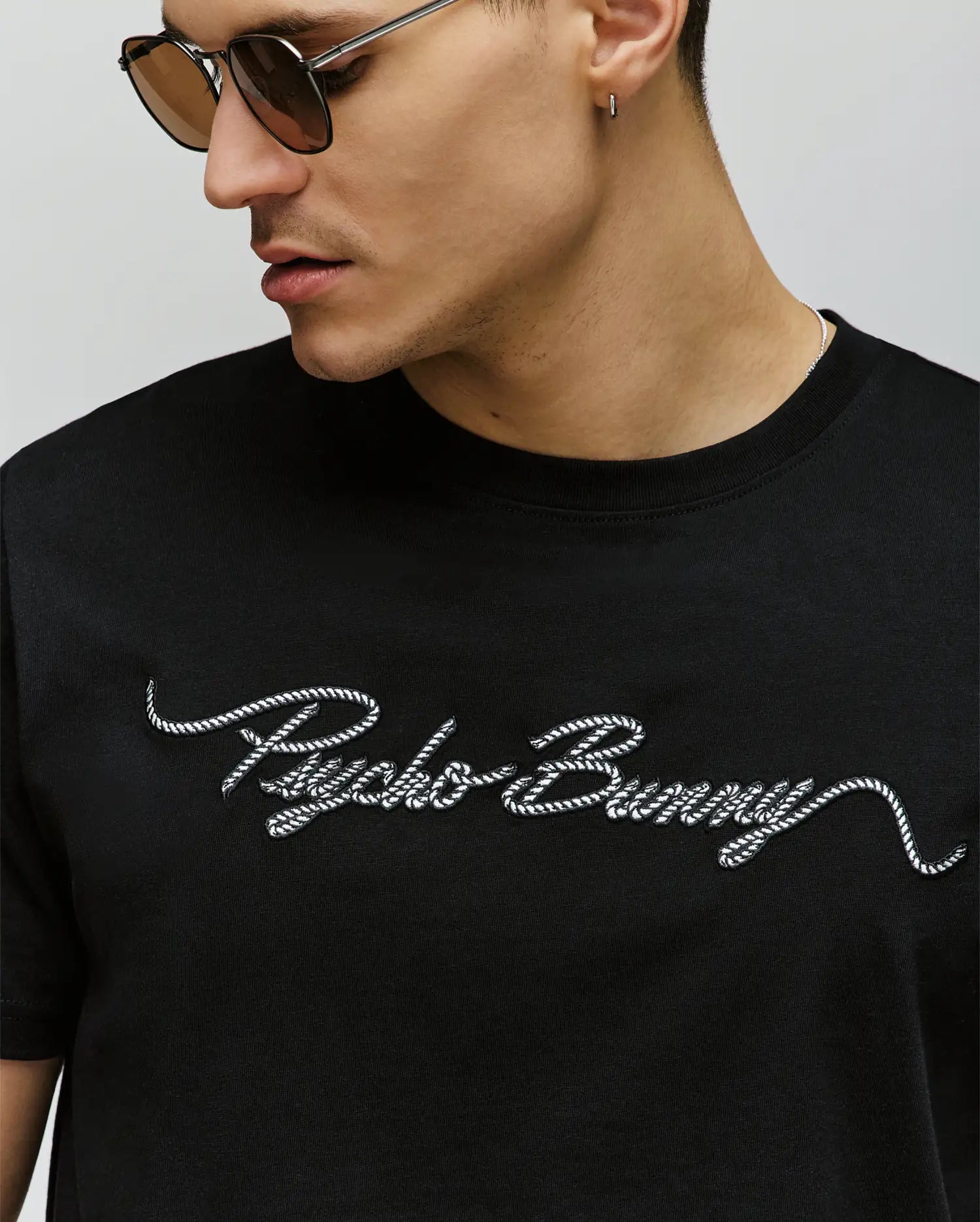 A man wearing sunglasses and a black Psycho Bunny MENS CYRUS WORDMARK GRAPHIC TEE (MT0200018) with white script "Psycho Bunny" embroidered across the chest.