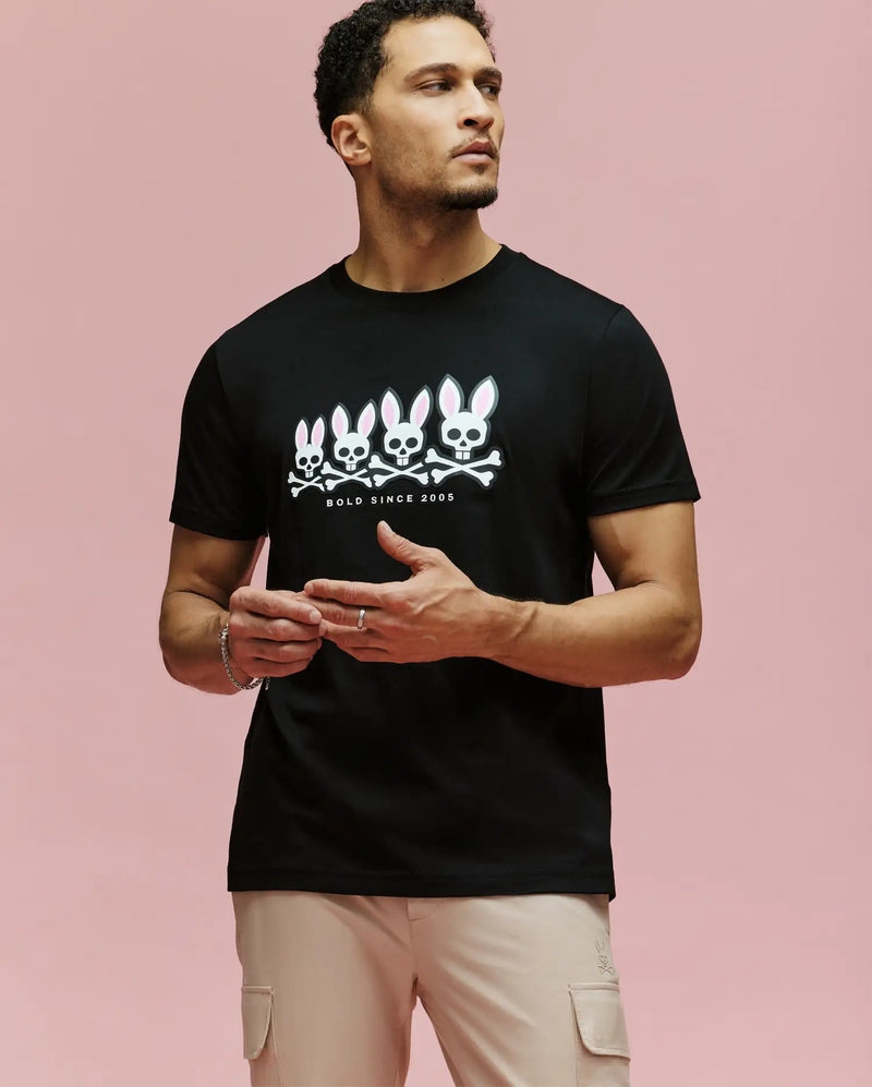 A man models the Psycho Bunny MENS ARCHIVE GRADIANT BUNNY GRAPHIC TEE (MT0200029), a bold black shirt with four Bunny skull-and-crossbones designs and "GOLD SINCE 2005" text, standing against a pink background, gesturing as he looks aside.