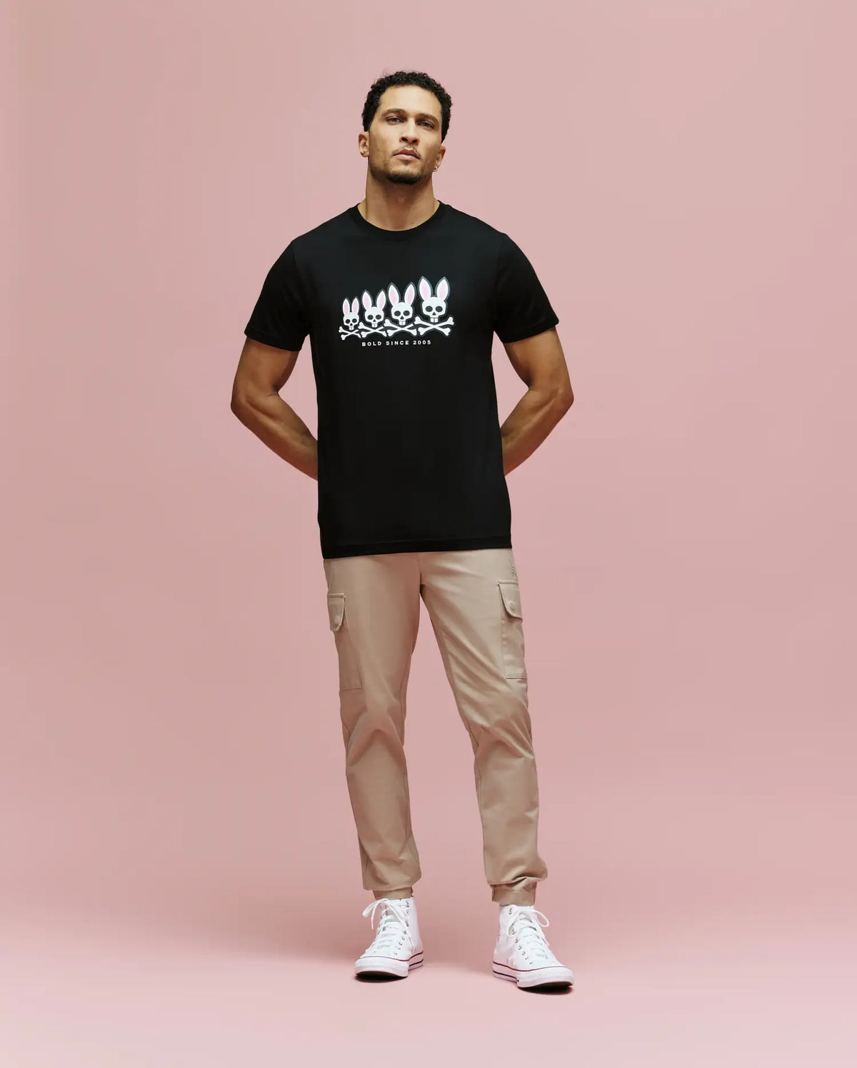 A man stands against a pink background wearing the Psycho Bunny MENS ARCHIVE GRADIANT BUNNY GRAPHIC TEE (MT0200029), paired with beige cargo pants and white high-top sneakers. He faces forward with his hands in his pockets.