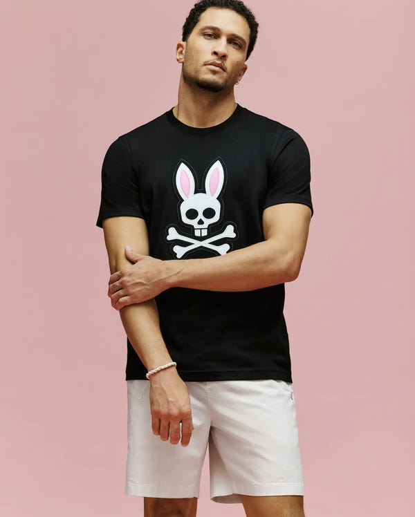 A man stands against a pink background wearing the Psycho Bunny MENS ARCHIVE BUNNY GRAPHIC TEE (MT0200031), a regular fit black T-shirt with an embroidered Bunny skull and crossbones, paired with light-colored shorts. He looks at the camera, one arm crossed.