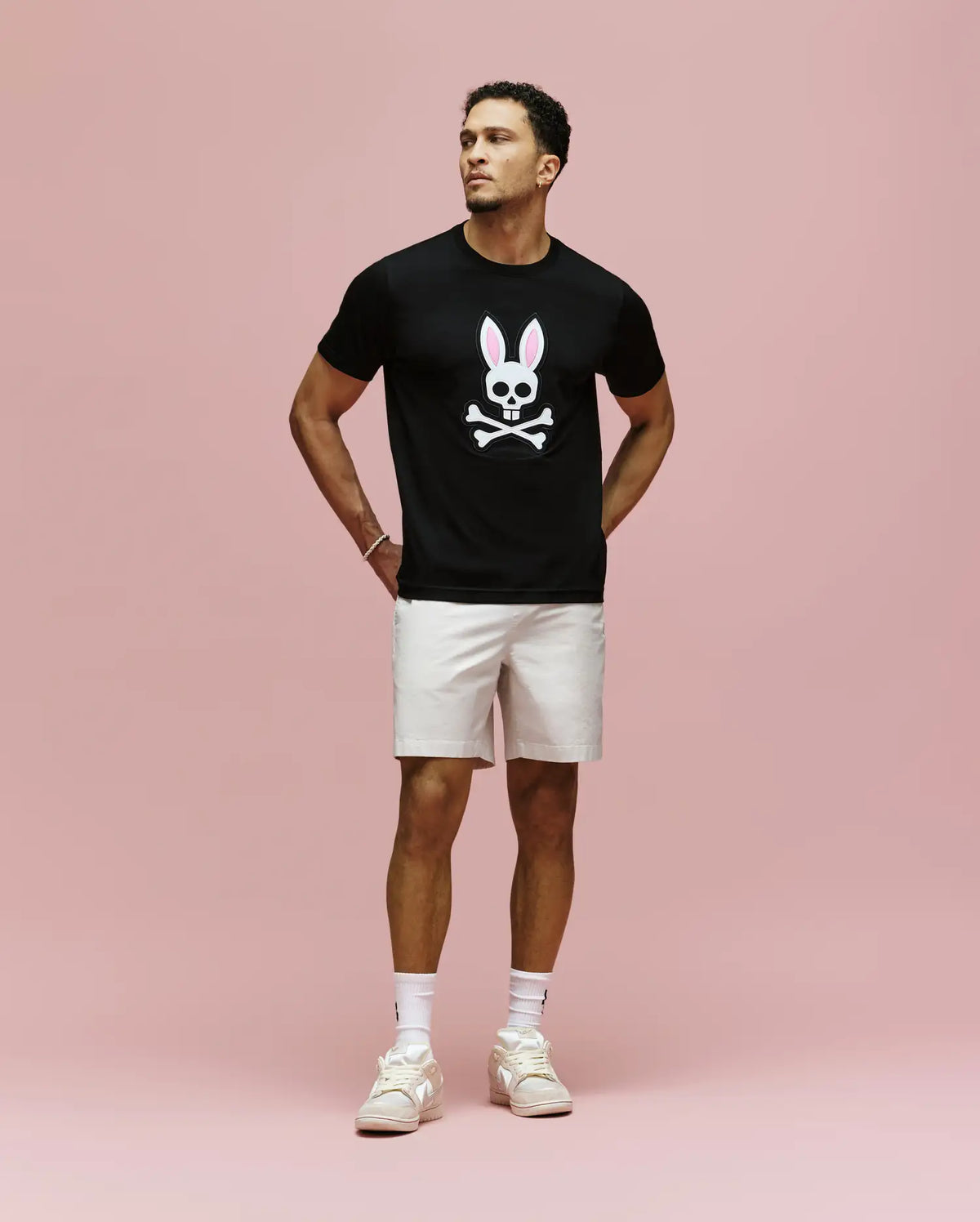 A man stands against a pink background wearing the Psycho Bunny MENS ARCHIVE BUNNY GRAPHIC TEE (MT0200031), black with an embroidered bunny and crossbones, paired with light shorts, white socks, and sneakers. Hands behind his back, he gazes to the side.