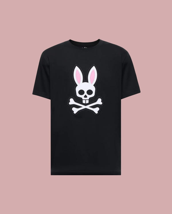 The Psycho Bunny MENS ARCHIVE BUNNY GRAPHIC TEE (MT0200031) is a black Pima cotton regular fit tee with a large white skull, pink bunny ears, and crossed bones on the front, shown on a light pink background for standout style and comfort.