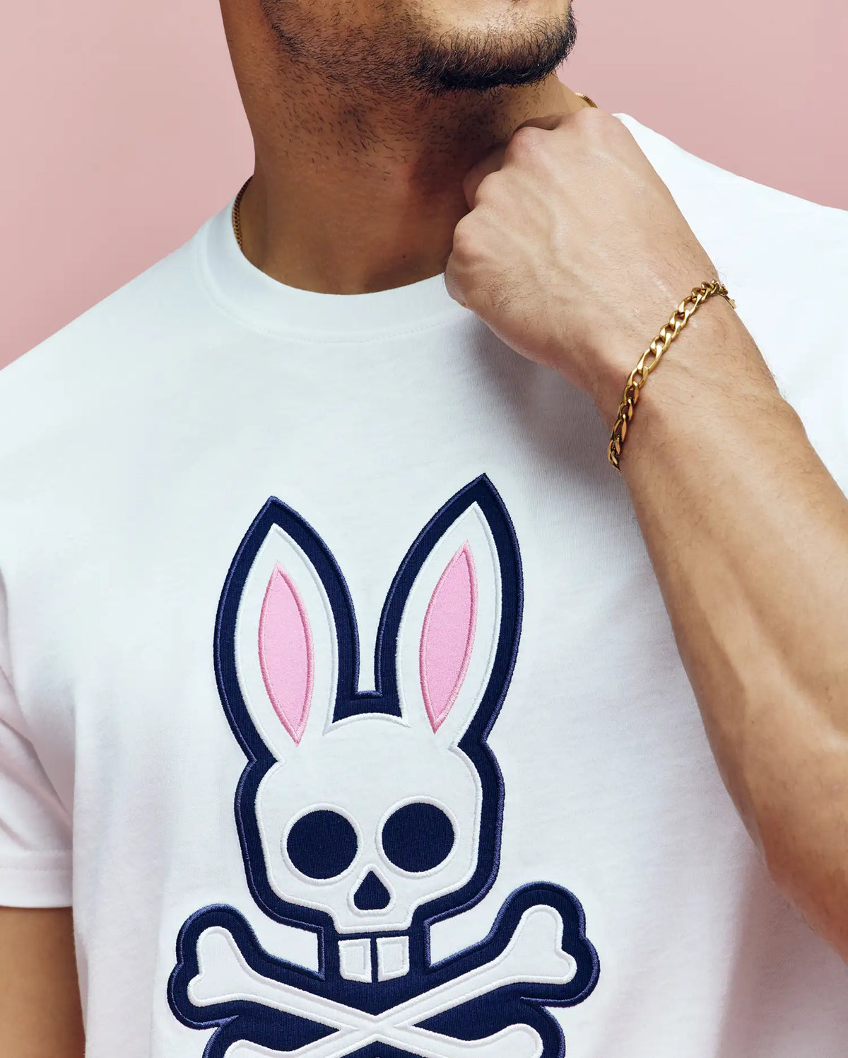 A person wearing the Psycho Bunny MENS ARCHIVE BUNNY GRAPHIC TEE (MT0200031), featuring a white Pima cotton shirt with a large skull and crossbones bunny ears graphic, gold chain bracelet, hand on chin, posed against a pink background.