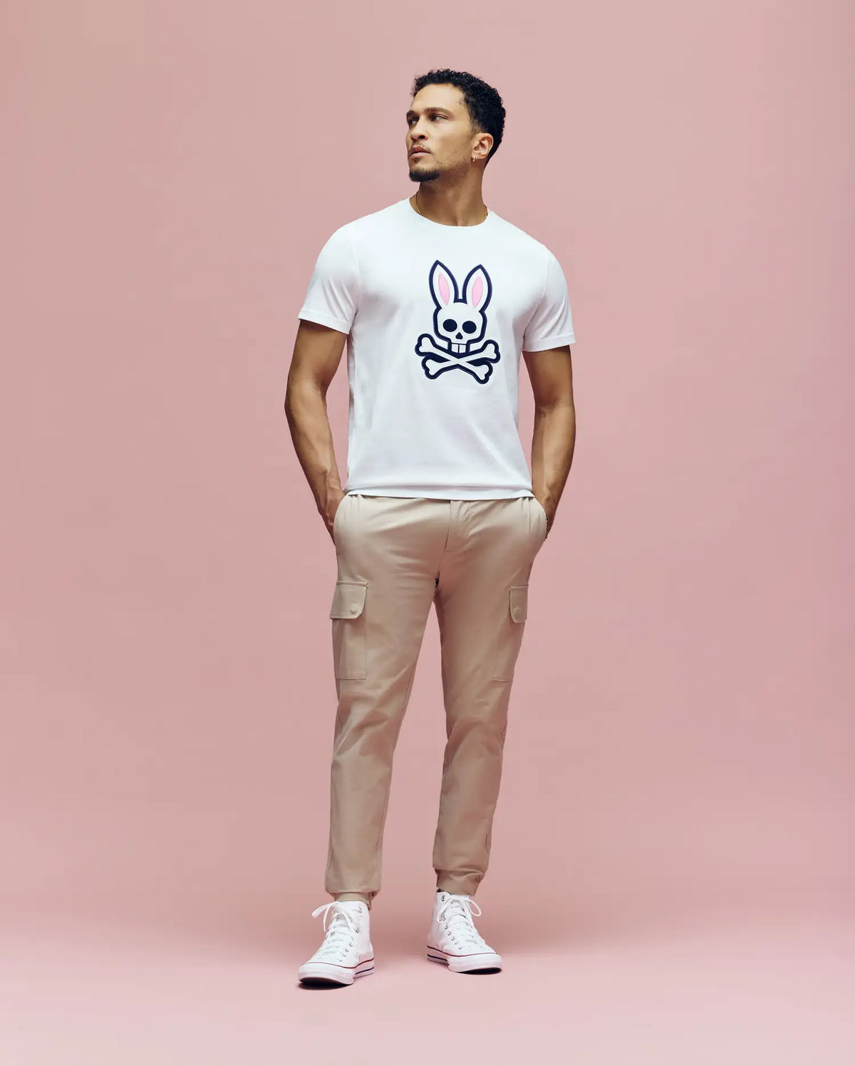 A man stands against a pink background, wearing the Psycho Bunny MENS ARCHIVE BUNNY GRAPHIC TEE (MT0200031) in white, with an embroidered bunny and crossbones graphic, paired with beige cargo pants and white sneakers. He looks up with his hands in his pockets.