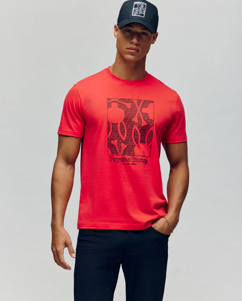 A man in a black cap, dark pants, and the Psycho Bunny MENS GLENN GRAPHIC TEE - MT0200034 with abstract patterns stands against a plain light gray background.
