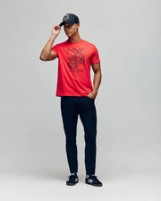 A man poses against a plain background wearing a black cap, black pants, black Adidas sneakers with white stripes, and the Psycho Bunny MENS GLENN GRAPHIC TEE - MT0200034 in red featuring a bold bunny graphic. He holds his cap and has one hand in his pocket.