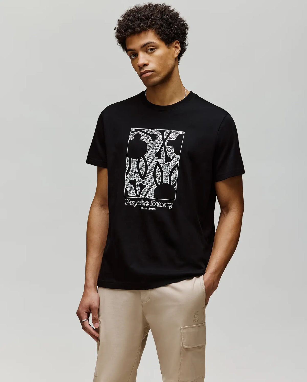 A man wearing the Psycho Bunny MENS GLENN GRAPHIC TEE (MT0200034), a black Pima cotton t-shirt with a Bunny graphic, stands against a plain light background, his hands in the pockets of his beige pants.
