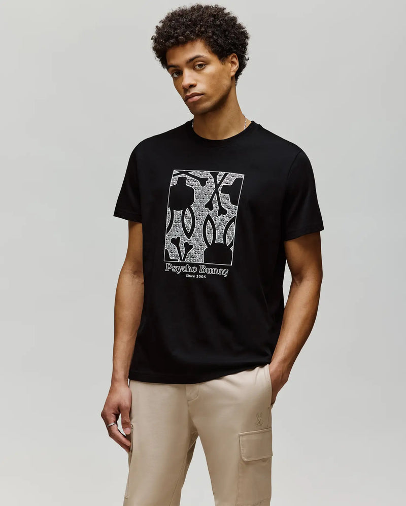 A man wearing the Psycho Bunny MENS GLENN GRAPHIC TEE (MT0200034), a black Pima cotton t-shirt with a Bunny graphic, stands against a plain light background, his hands in the pockets of his beige pants.